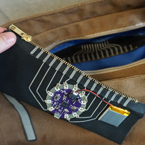 This image shows a preview of the rapid iron-on project. More specifically, a zip-on component with an Arduino Lilypad is shown that was unzipped from a smart messenger bag that is visible in the background.